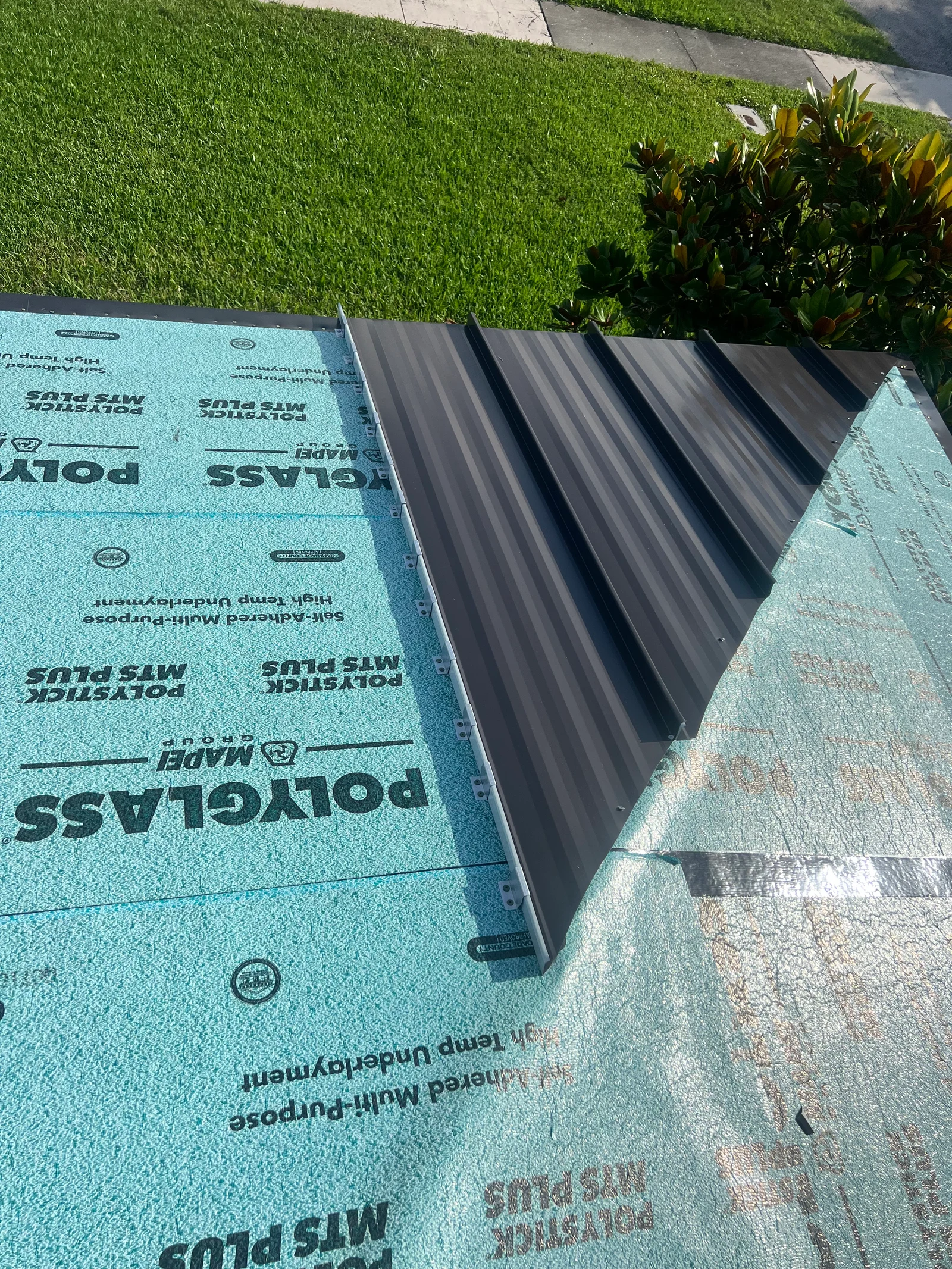 Dark standing-seam metal panels with exposed edge clips over blue Polystick MTS Plus underlayment.