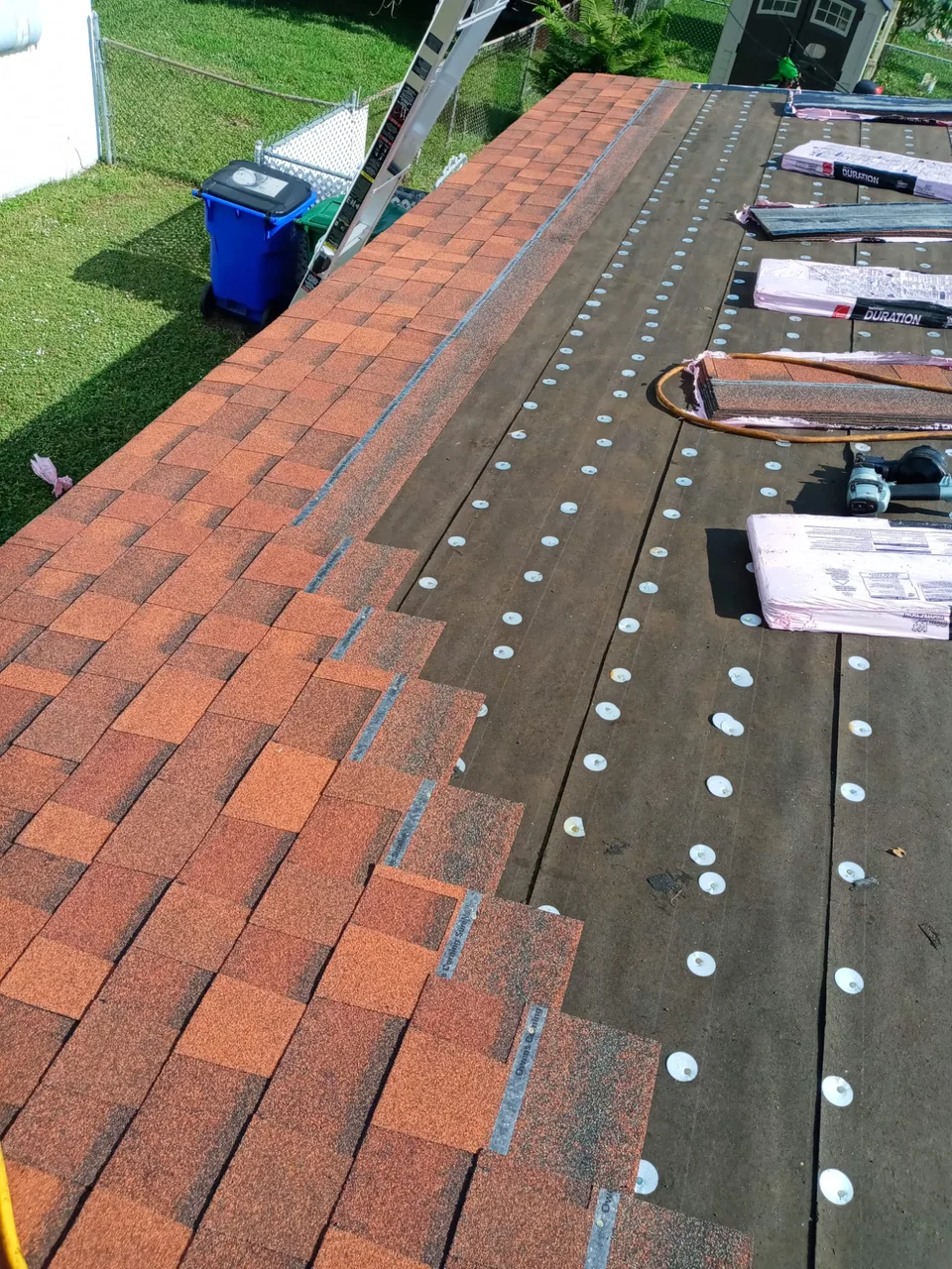 Reddish-brown shingles being installed along a roof edge beside capped underlayment and shingle bundles.